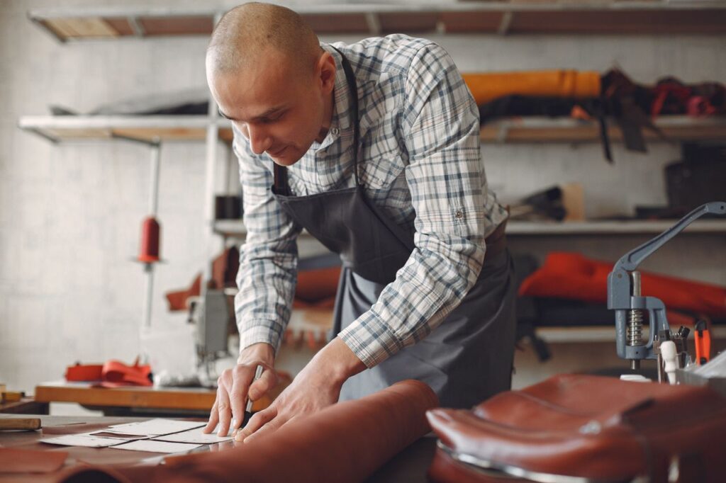 Concentrated artisan in plaid shirt and apron lining piece of leather with ruler and marker while working in professional workshop