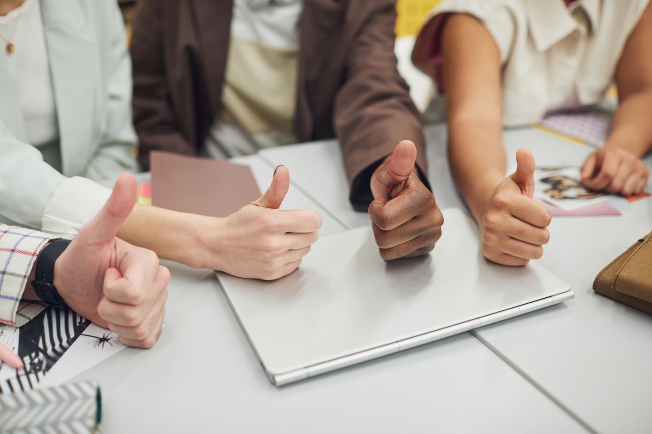 Four diverse hands showing thumbs up over a laptop on a desk, symbolizing teamwork and approval.